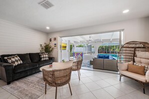Ein modernes Wohnzimmer mit einer Couch, einem Esstisch und Stühlen. Ein großes Fenster bietet Blick auf einen Swimmingpool.