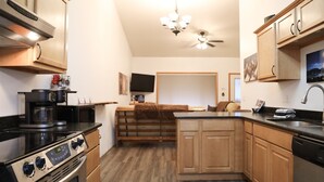 A kitchen with a stove, oven, and coffee maker. There is a dining area with a bench and a television. The kitchen has wooden cabinets and a black countertop. - Serene Morning Meadow Condo  #93, at Wallowa Lake (Joseph)