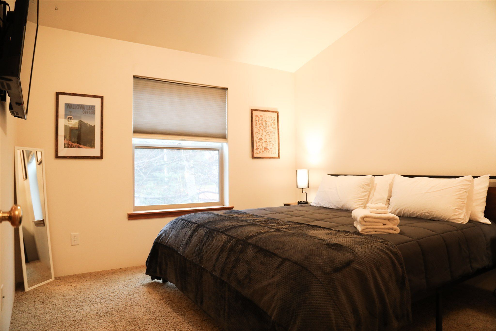 A bedroom with a bed, a window with blinds, a bedside lamp, and two framed pictures on the wall.