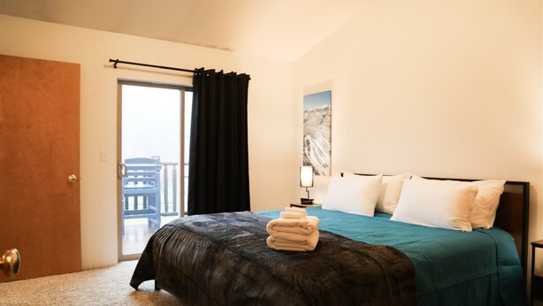 A hotel room with a bed, a nightstand, a lamp, a window with curtains, and a door. - Serene Morning Meadow Condo  #93, at Wallowa Lake (Joseph)