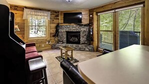 A cozy cabin interior with a stone fireplace, wooden walls, a television mounted above it, and a large window with blinds. - Affordable Pigeon Forge Cabin 1 Mile to Strip! (Pigeon Forge)