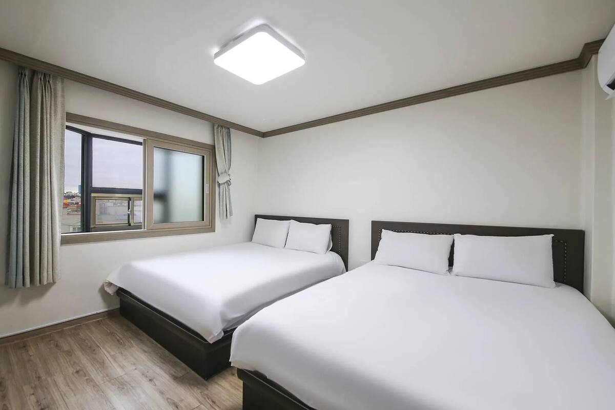 Une chambre d’hôtel avec deux lits simples, une fenêtre avec des rideaux, un luminaire au plafond et un bureau avec un sèche-cheveux bleu.