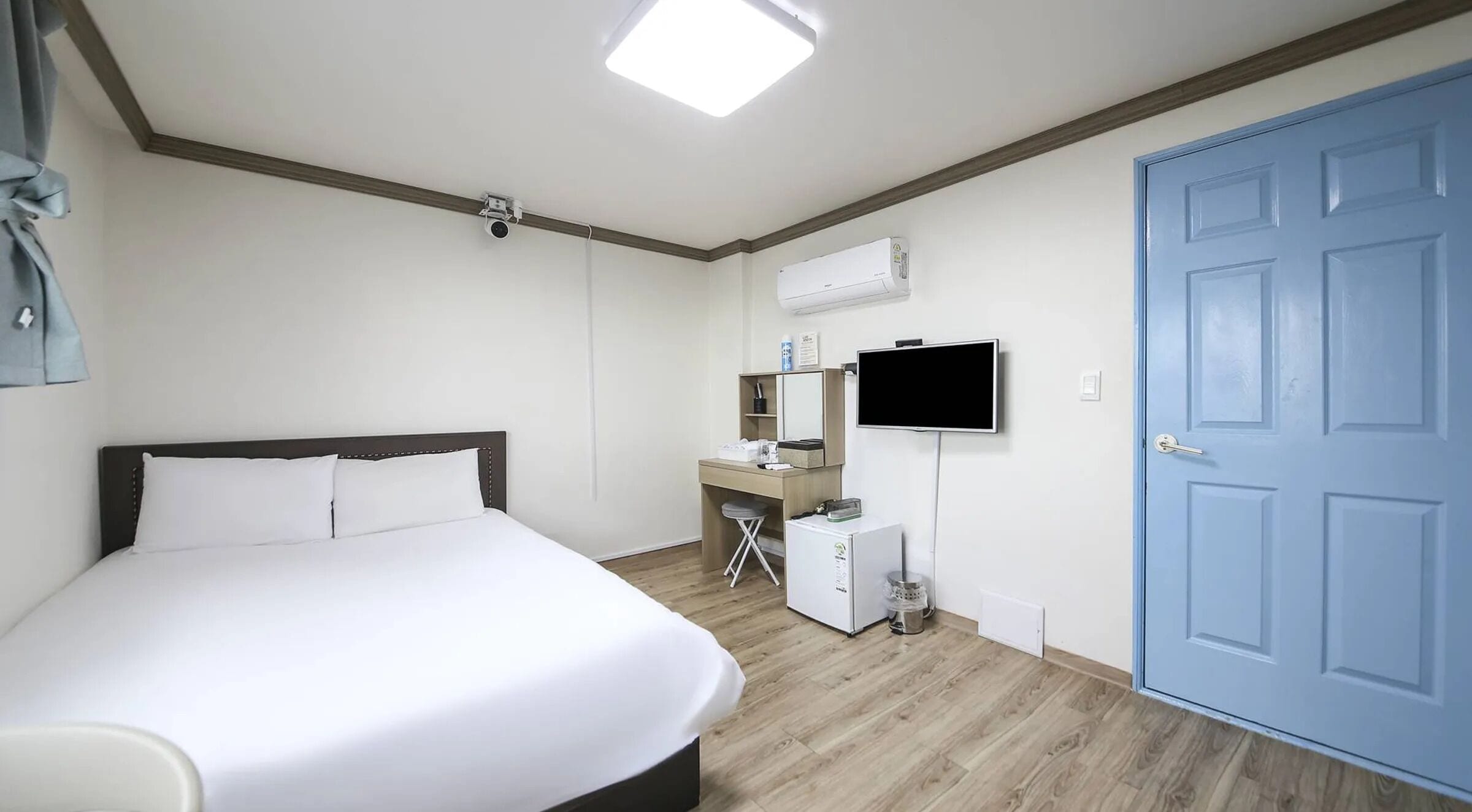 A hotel room with a bed, a desk with a chair, a television, and a blue door.