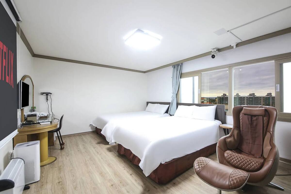 Une chambre d’hôtel avec un grand lit, un bureau équipé d’un ordinateur, une chaise et une vue sur la ville par une grande fenêtre.