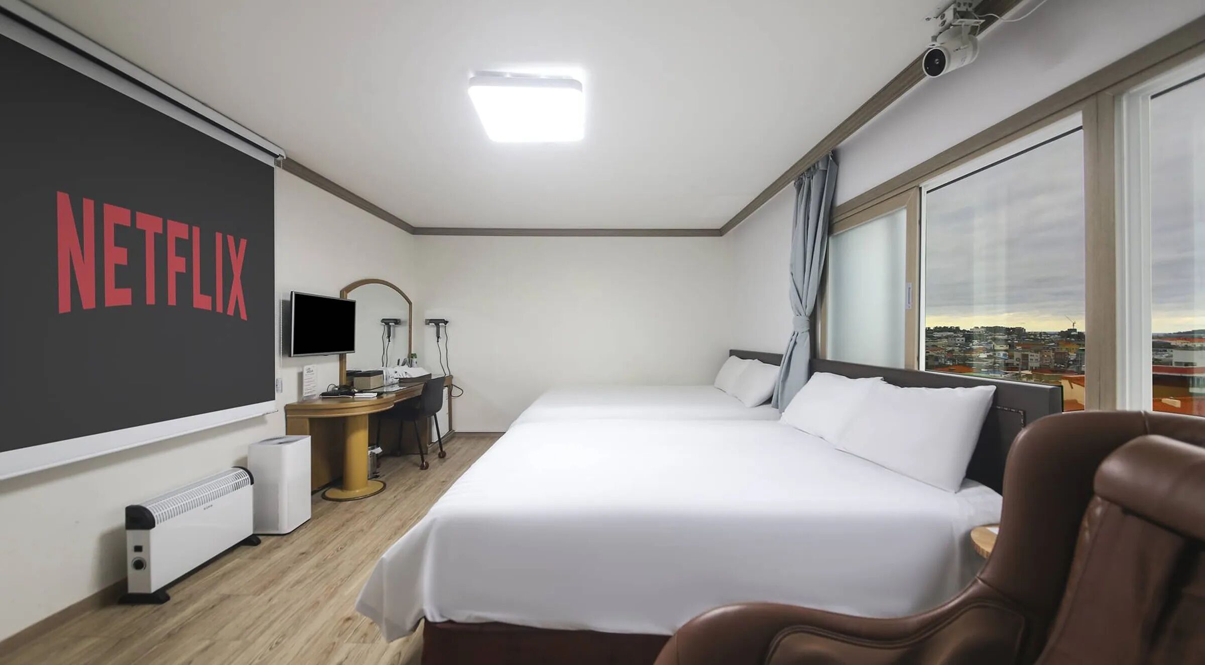 Habitación de hotel con una cama grande, un escritorio con computadora, una silla, un televisor montado en la pared y vistas a la ciudad a través de la ventana.