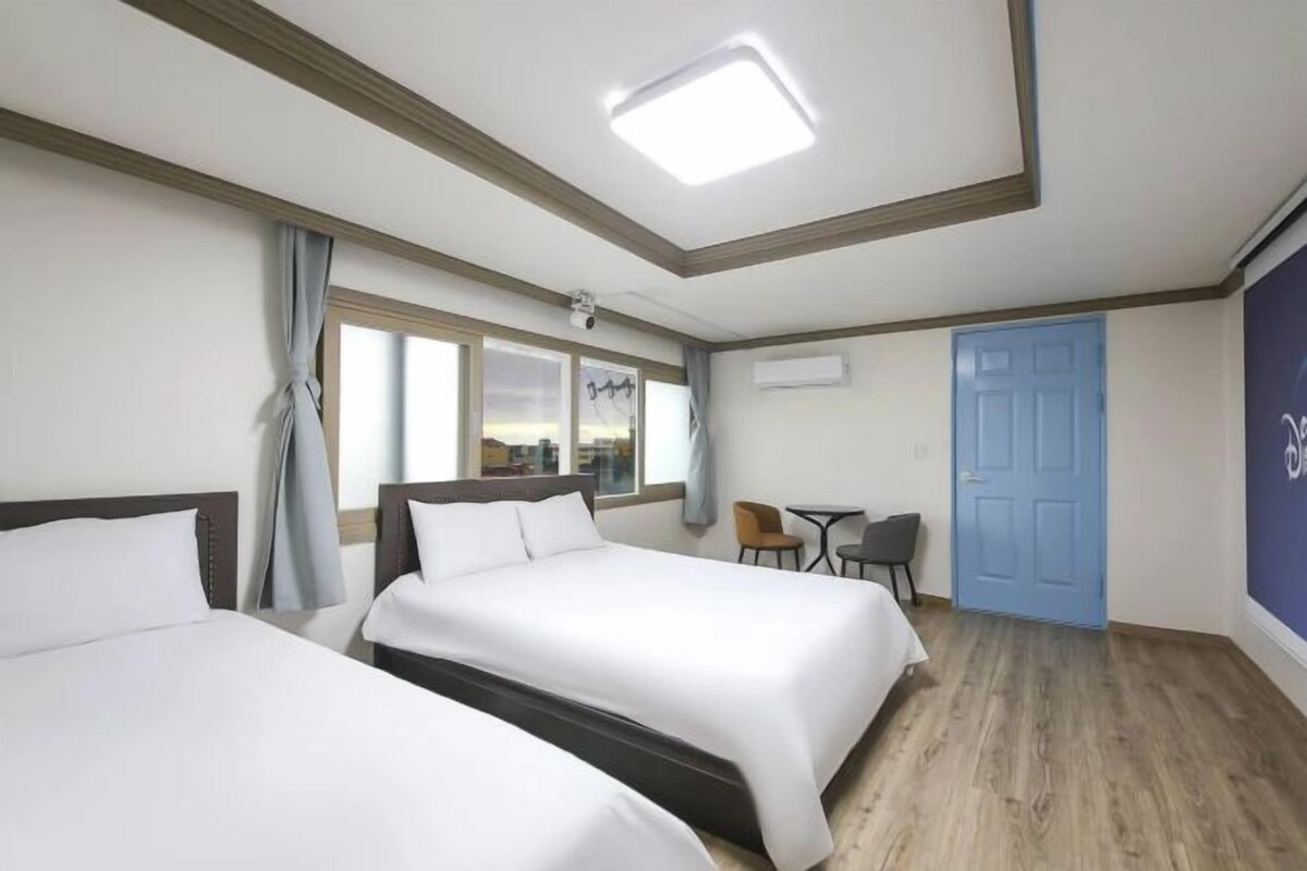 Une chambre d’hôtel avec deux lits, un bureau, une chaise, une fenêtre avec des rideaux, une porte et un écran de projection.