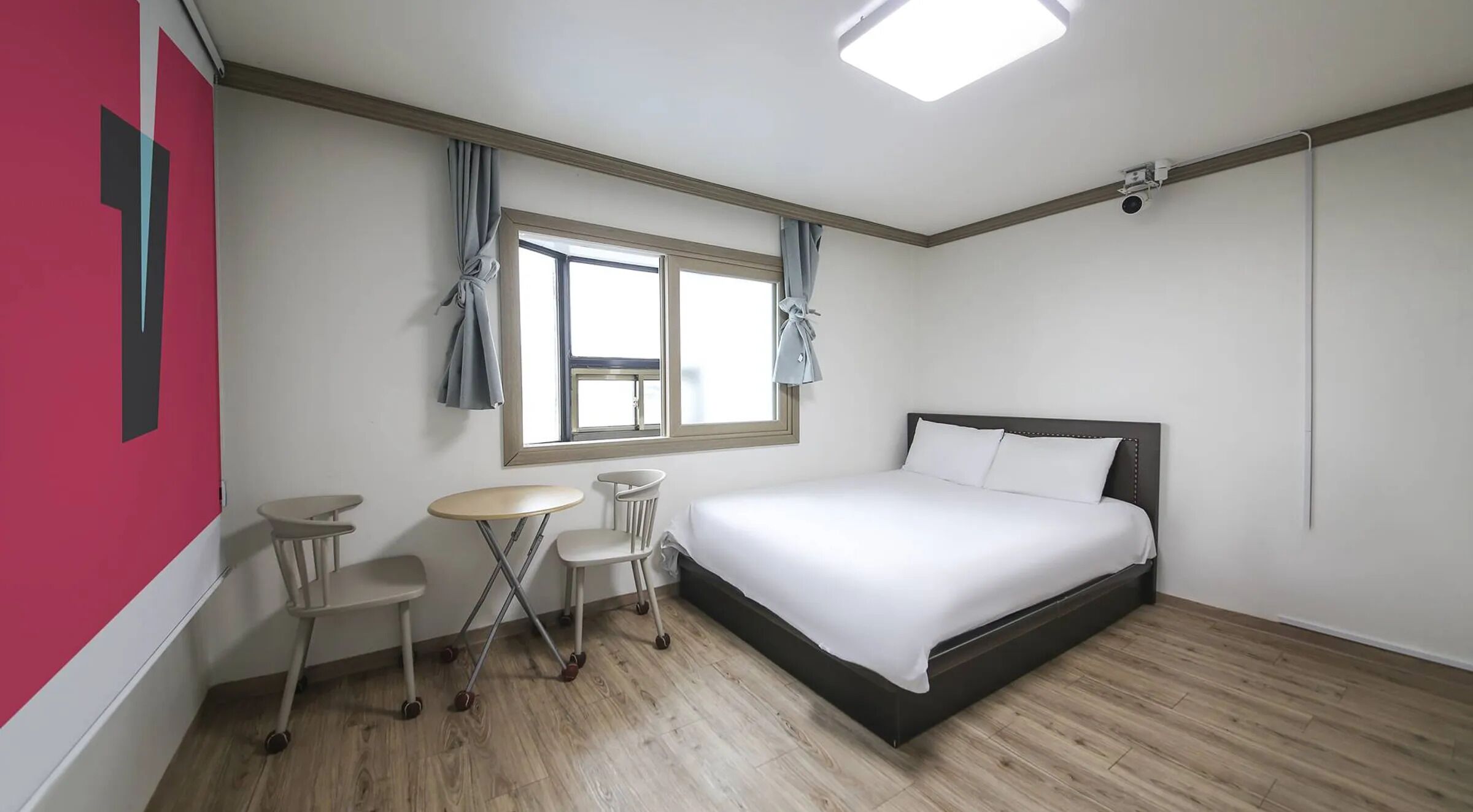 A hotel room with a bed, a small table, two chairs, a window with curtains, and a ceiling light.