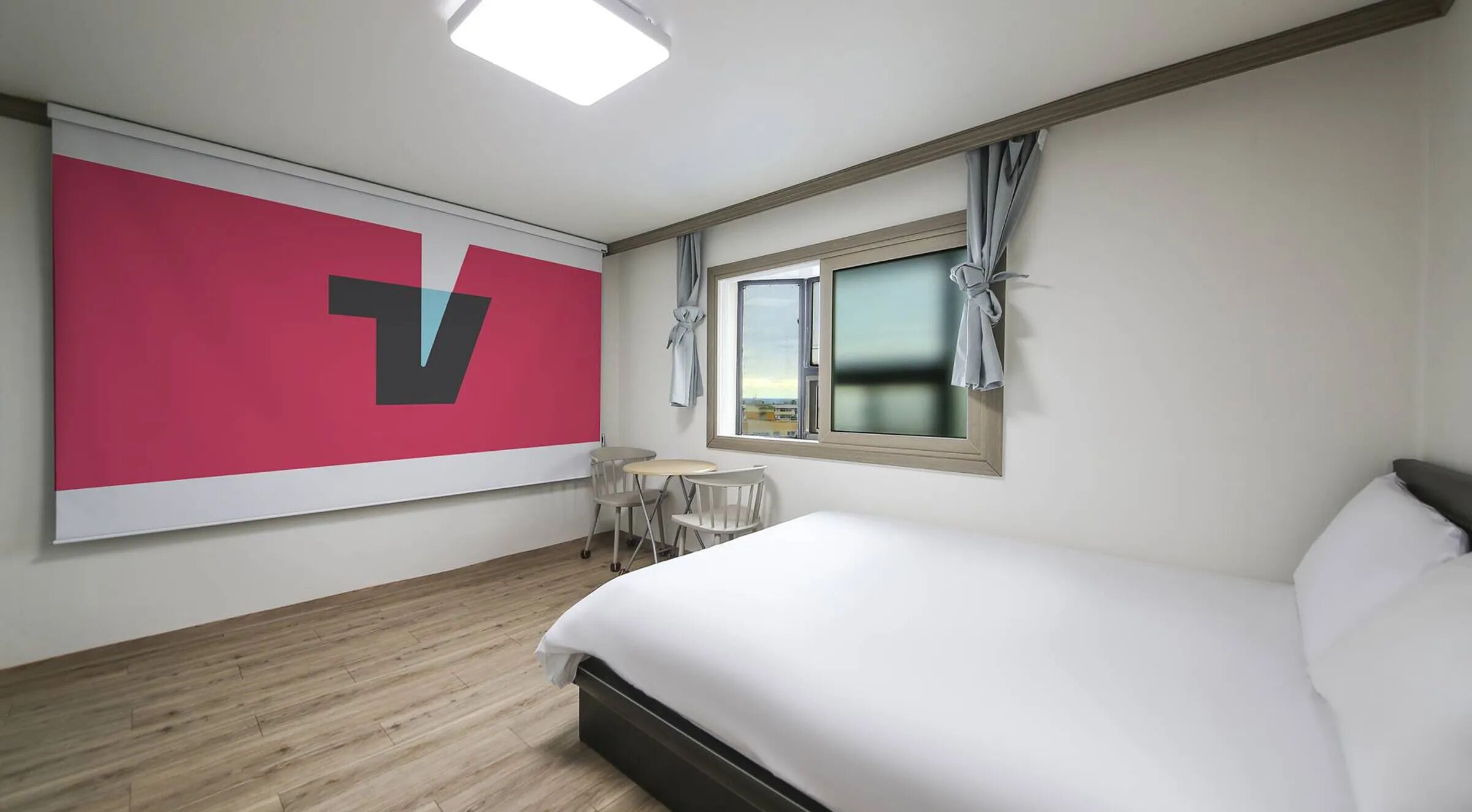 Une chambre d’hôtel avec un lit, une petite table, une chaise, une fenêtre avec des rideaux et un grand élément décoratif mural.