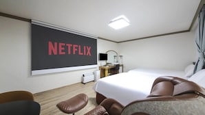 A hotel room with a large bed, a desk with a computer, a chair, a TV screen displaying "NETFLIX," and a bedside table with a lamp. - Top Island Hotel (Jeju City)