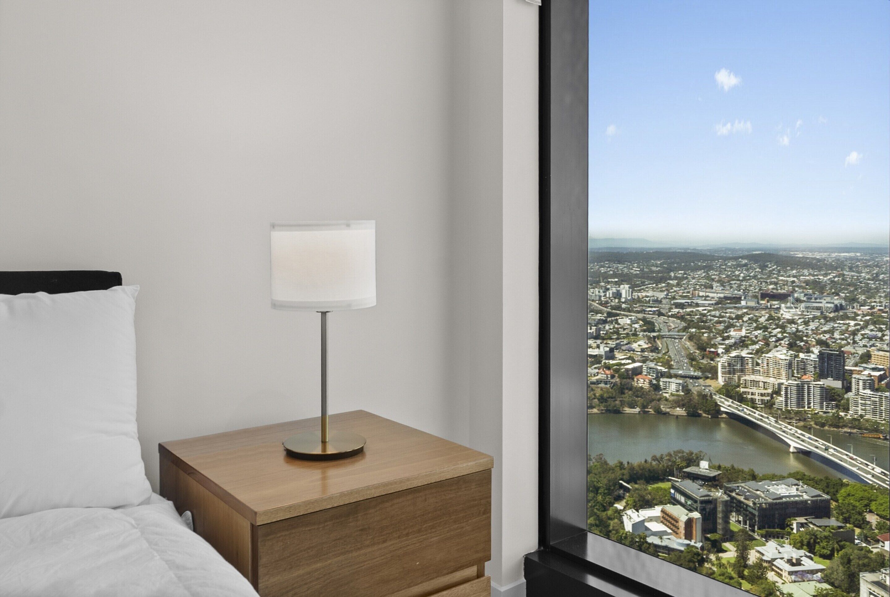 A bedroom with a bed, a bedside table with a lamp, and a large window offering a city view.