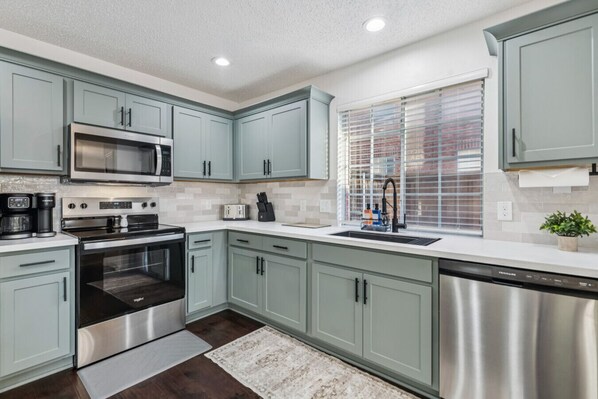 A modern kitchen with stainless steel appliances, light blue cabinetry, and a window with blinds. - Billiards & Blue Water (Grand Prairie)