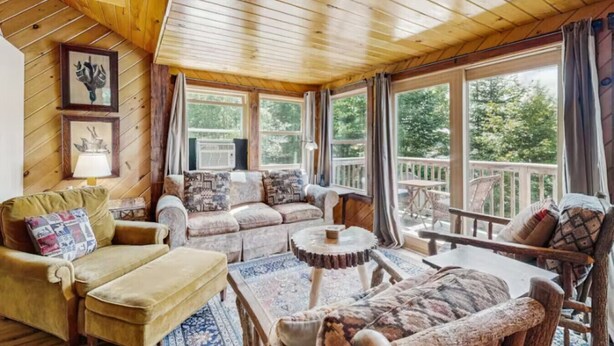 A cozy cabin interior with wooden ceilings, a sofa set, a coffee table, and large windows offering a view of the outdoors.