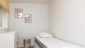 Une petite pièce blanche, avec un lit, un radiateur, un petit tabouret en bois et deux tableaux encadrés accrochés au mur.