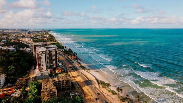 An aerial view of a coastal city with a beach, buildings, and a clear sky. - Apartment across from the beach for millionaires. (Ilhéus)