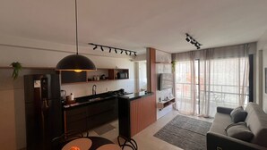 A modern kitchen with a central island, a dining area with a round table, and a living room with a sofa and a TV. - Apartment across from the beach for millionaires. (Ilhéus)
