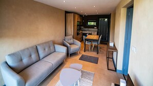 A modern living room with a sofa, armchairs, a dining table, and a kitchen area in the background. - Mountain-view Eco-friendly Cottage (Marcoux)