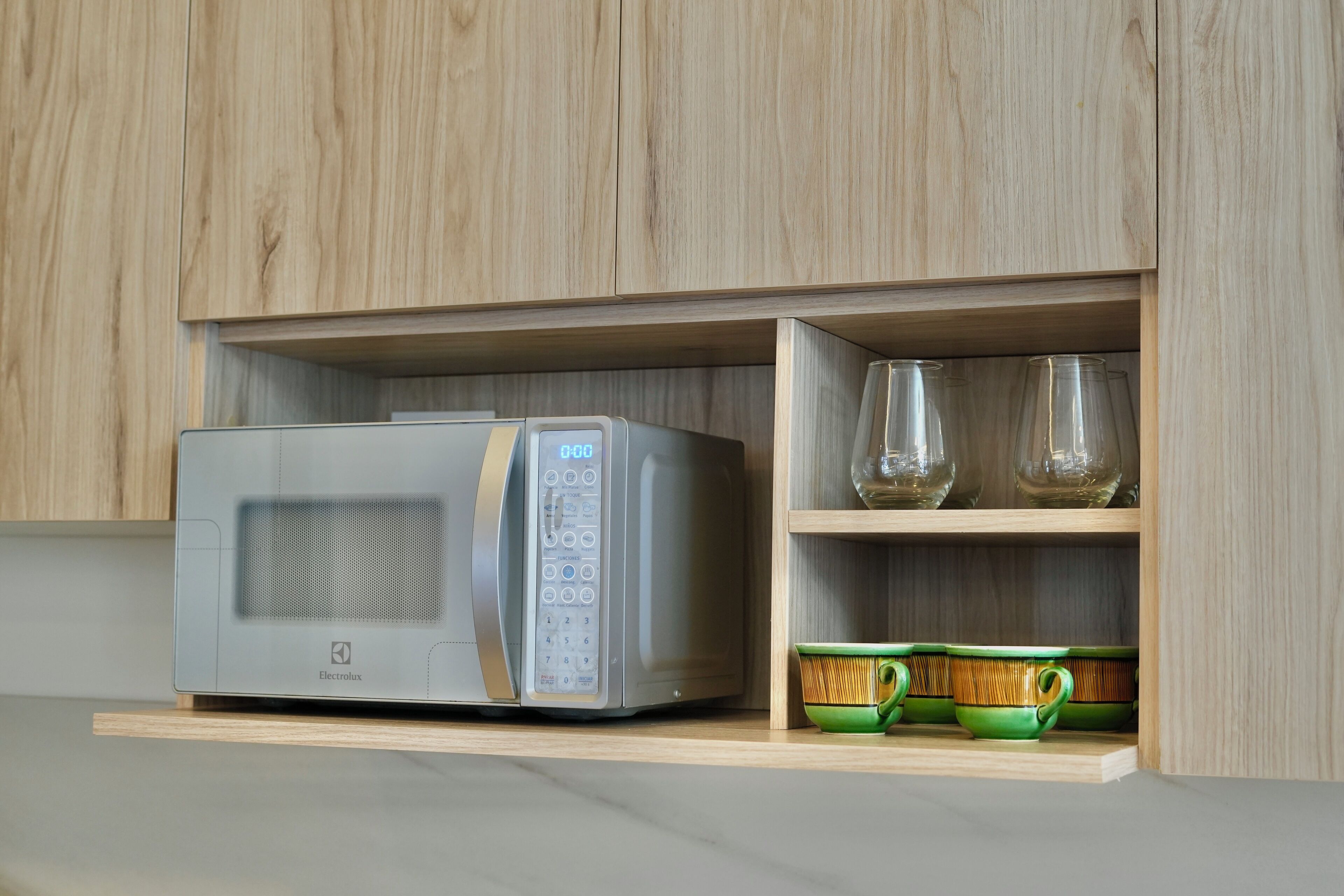 A microwave in a wooden cabinet with a shelf below containing glassware and green cups.