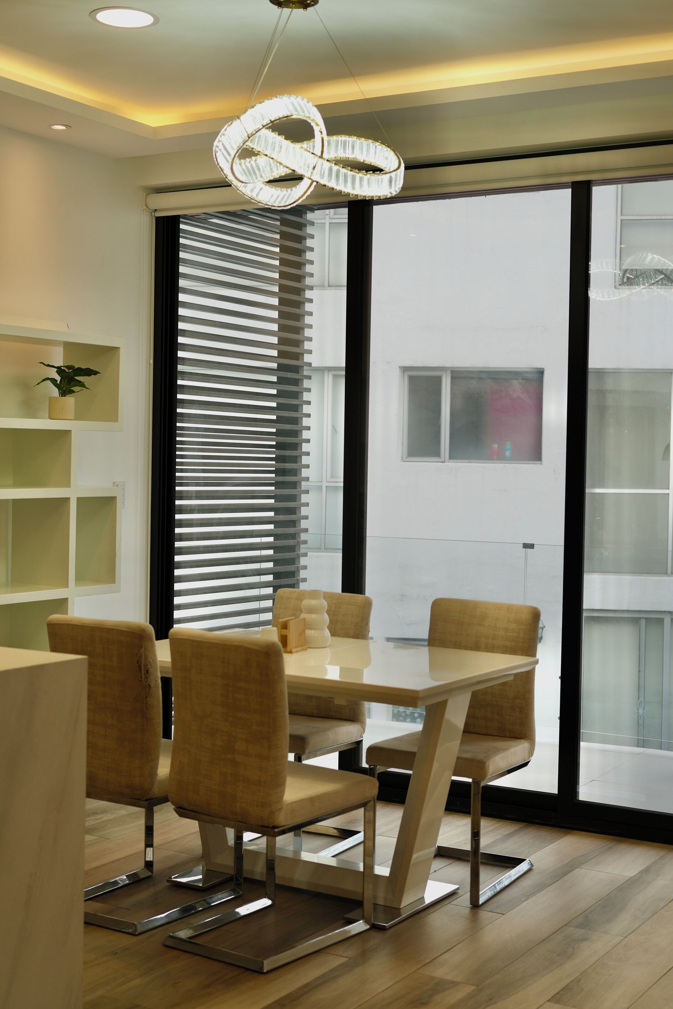 Un espace de salle à manger moderne avec une table et des chaises, de grandes fenêtres équipées de stores et un luminaire suspendu.