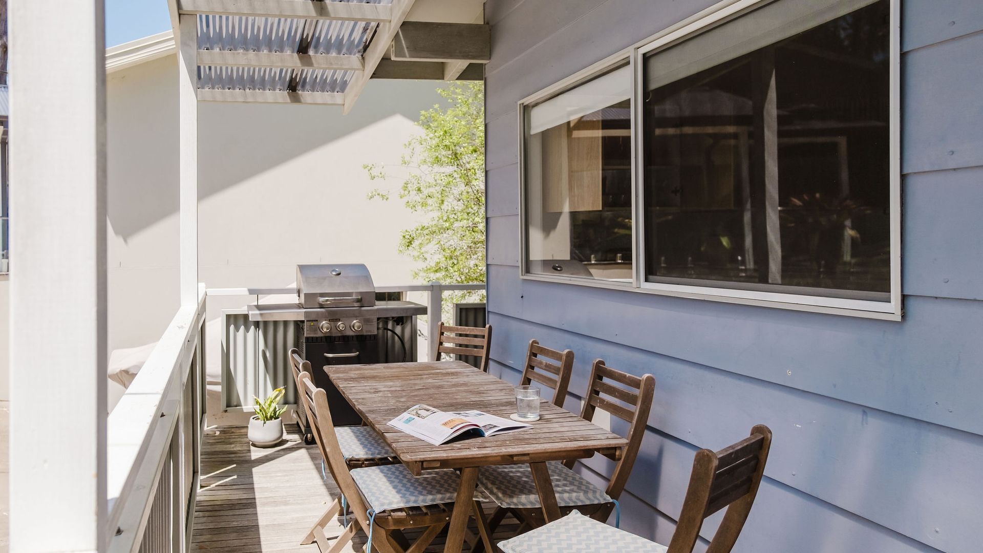 Eine Terrasse im Freien mit Holztisch und -stühlen, einem Grill und einem Fenster mit Blick auf den Garten.