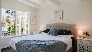 A bedroom with a large bed, a chandelier, a window with blinds, and a bedside table with a lamp and a vase of flowers. - Medona: Narrawallee Beach House (Narrawallee)