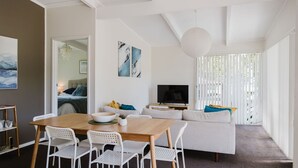 A modern living room with a wooden dining table, white chairs, a sofa, and a TV. - Medona: Narrawallee Beach House (Narrawallee)