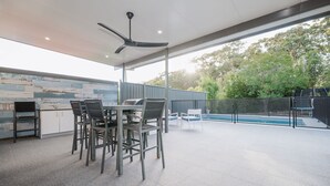Un patio al aire libre con una mesa, sillas, ventilador de techo, piscina y vistas a árboles.