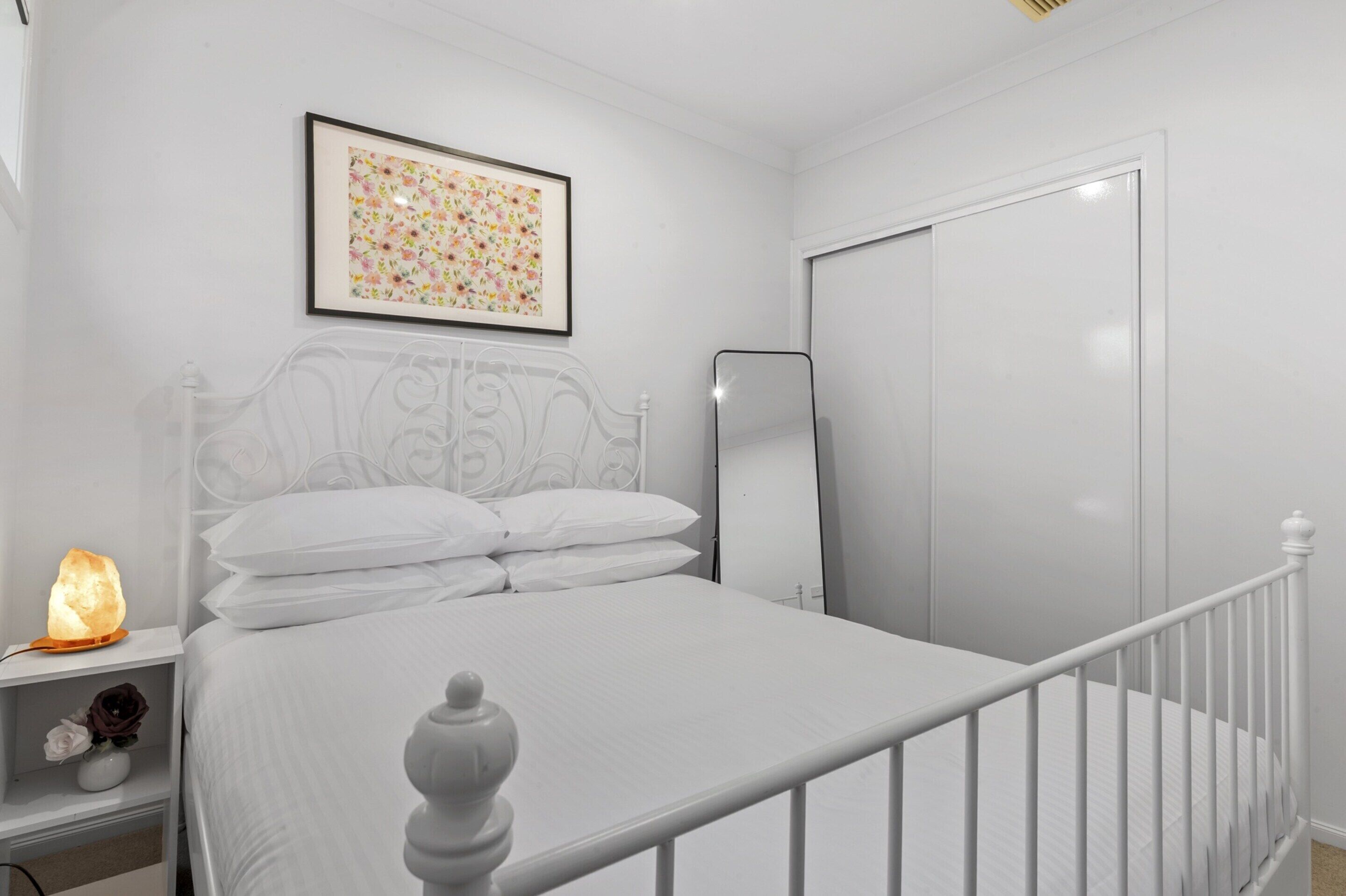 Een netjes opgemaakt bed met witte lakens en een hoofdbord, een nachtkastje met een zoutlamp, en een lijst met bloemkunst aan de muur.