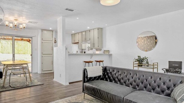 Una sala moderna con un sofá gris, un comedor con una mesa y sillas de madera, y una cocina con armarios blancos y un espejo redondo.
