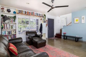 Ein Wohnzimmer mit Ledersofa, Bücherregal, Deckenventilator und Blick ins Freie.