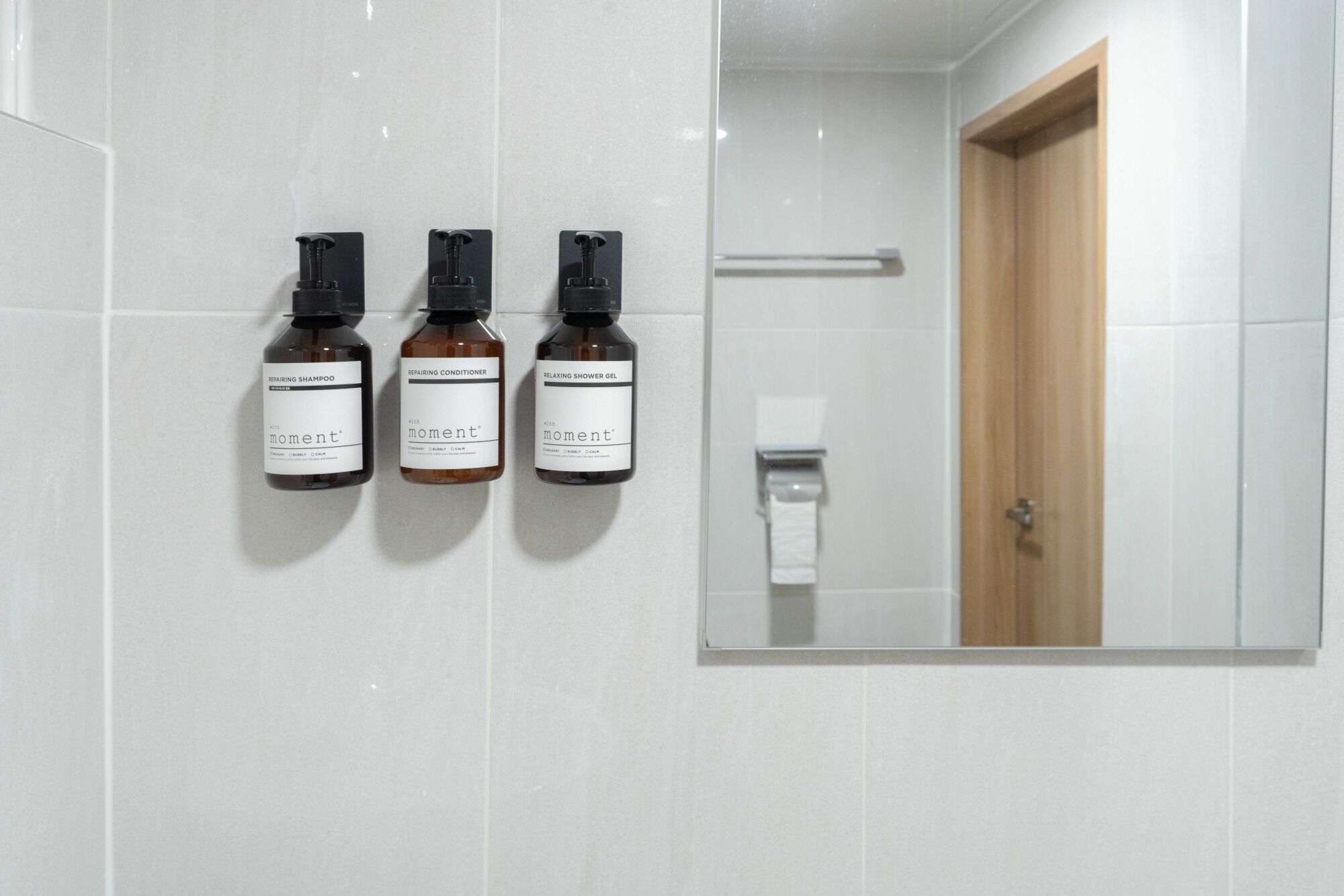 Un baño con tres botellas de productos 'moment' en la pared y una puerta de madera.