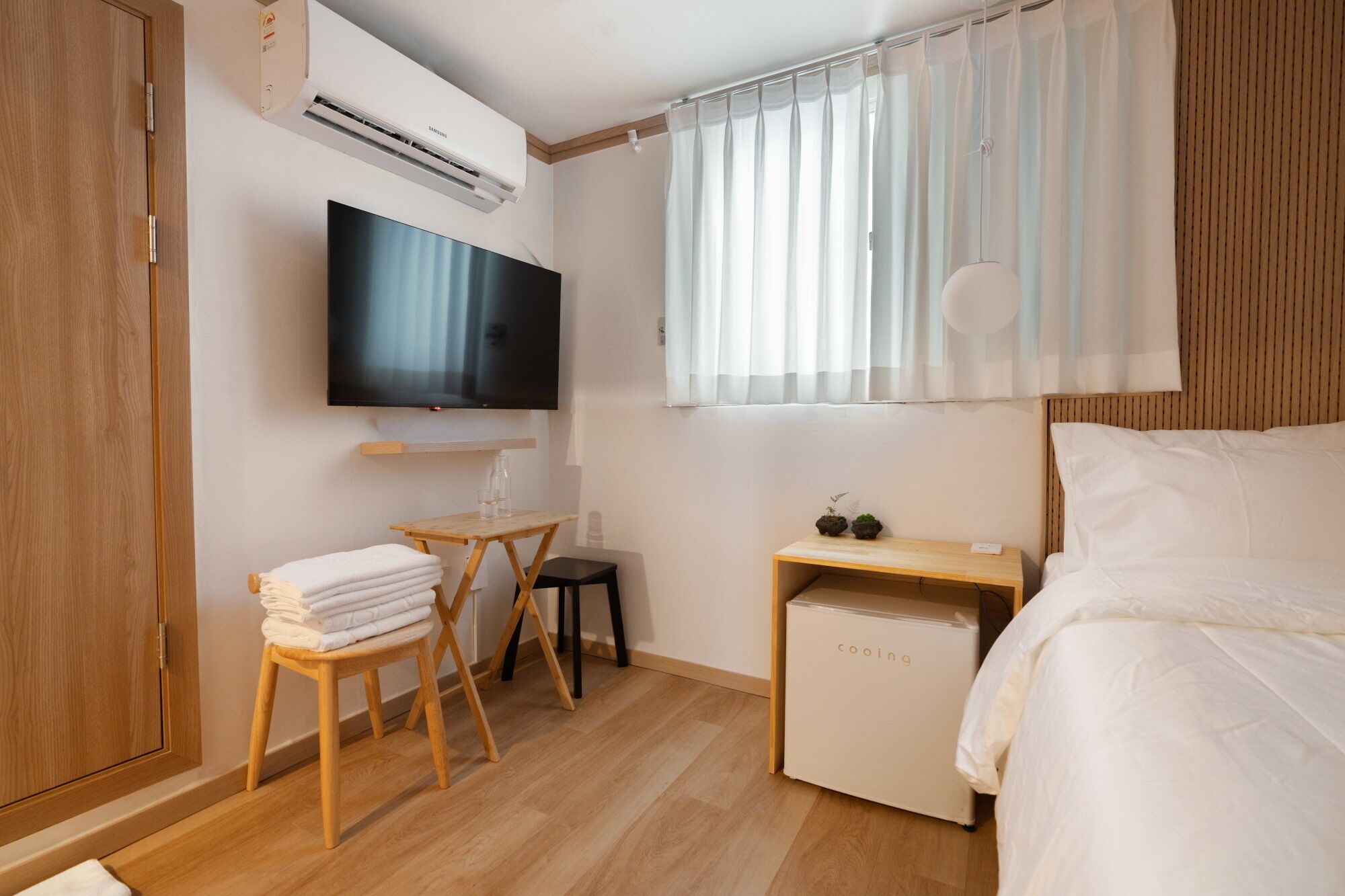 Een moderne hotelkamer met een bed, een televisie, een klein tafeltje met stoel, een minibar en een raam met dunne gordijnen.