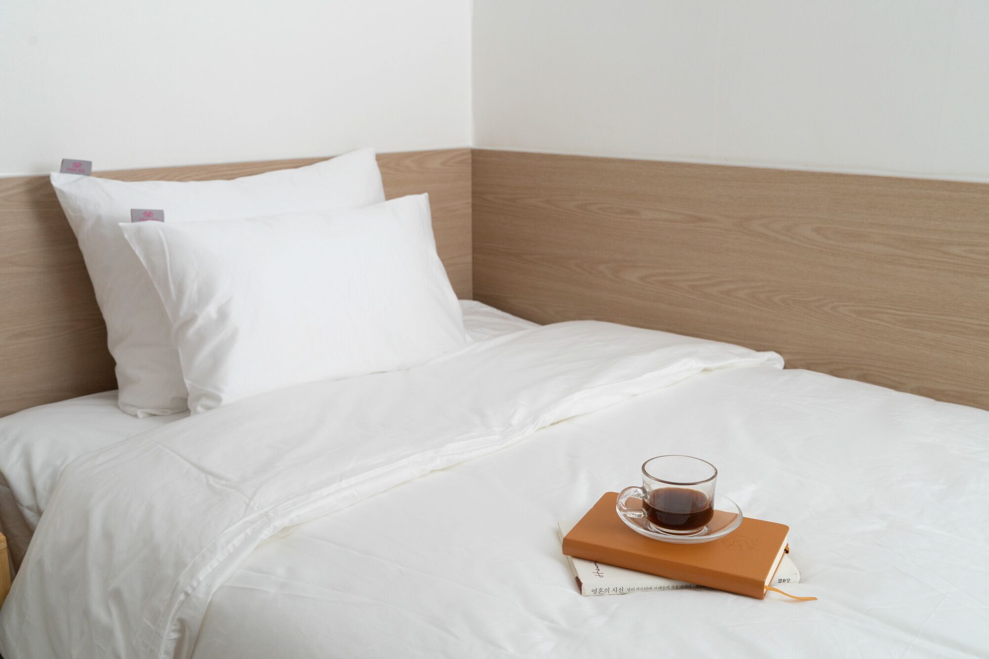 Una cama bien hecha con ropa blanca y almohadas, una taza de café sobre un libro y un cabecero de madera.