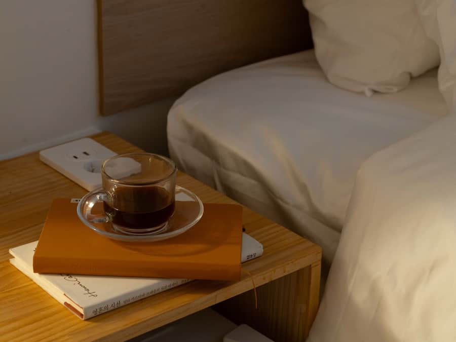 Una cama bien hecha con sábanas blancas, una mesita de noche con un libro y una taza de café, y un enchufe en la pared.