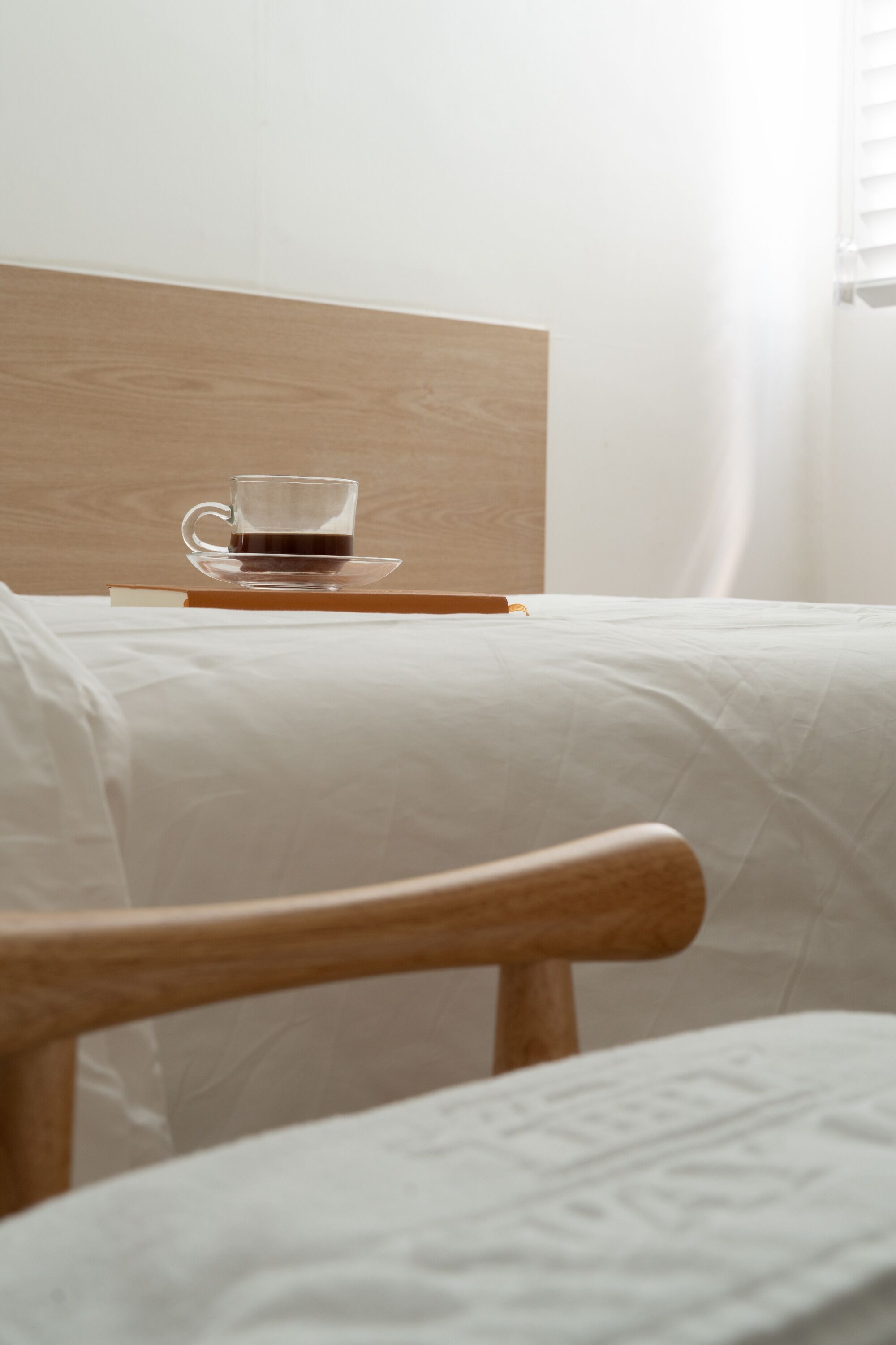 A neatly made bed with a wooden headboard, a cup of coffee on a saucer, and a wooden chair in the foreground.