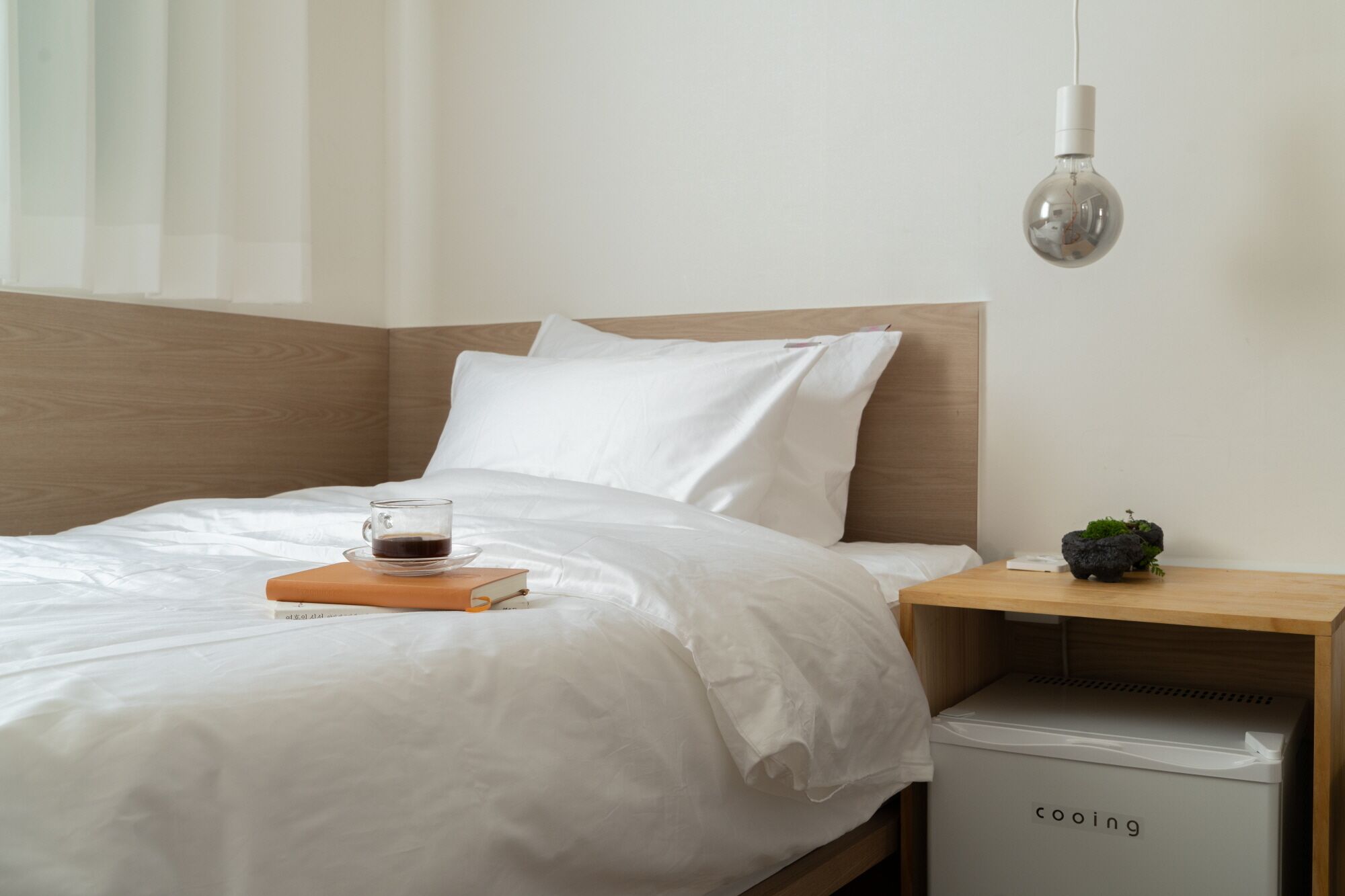 A neatly made bed with white linens, a glass of coffee on a book, a bedside table with a small plant, and a white appliance labeled "cooing" on the side.