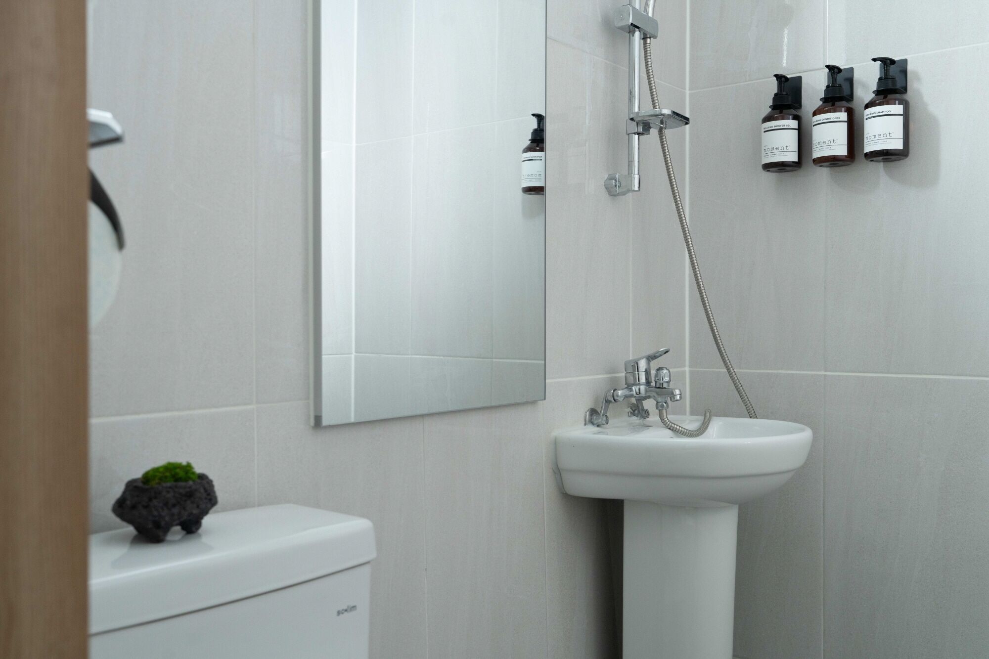 Un baño moderno con un lavabo blanco, un inodoro y una ducha. Hay tres botellas de champú en la pared.