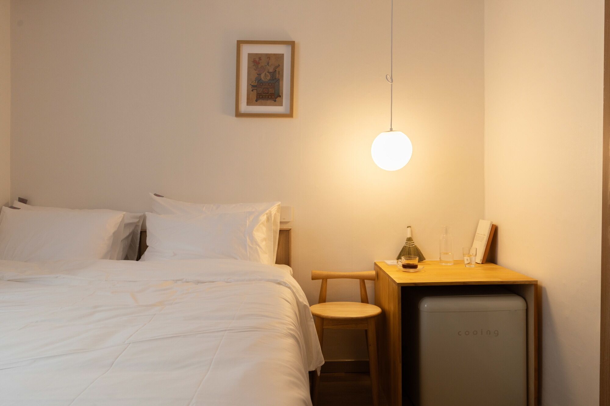 A hotel room with a bed, a bedside table with a lamp, a small stool, and a compact refrigerator.