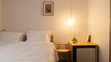 A hotel room with a bed, a bedside table with a lamp, a small stool, and a compact refrigerator.