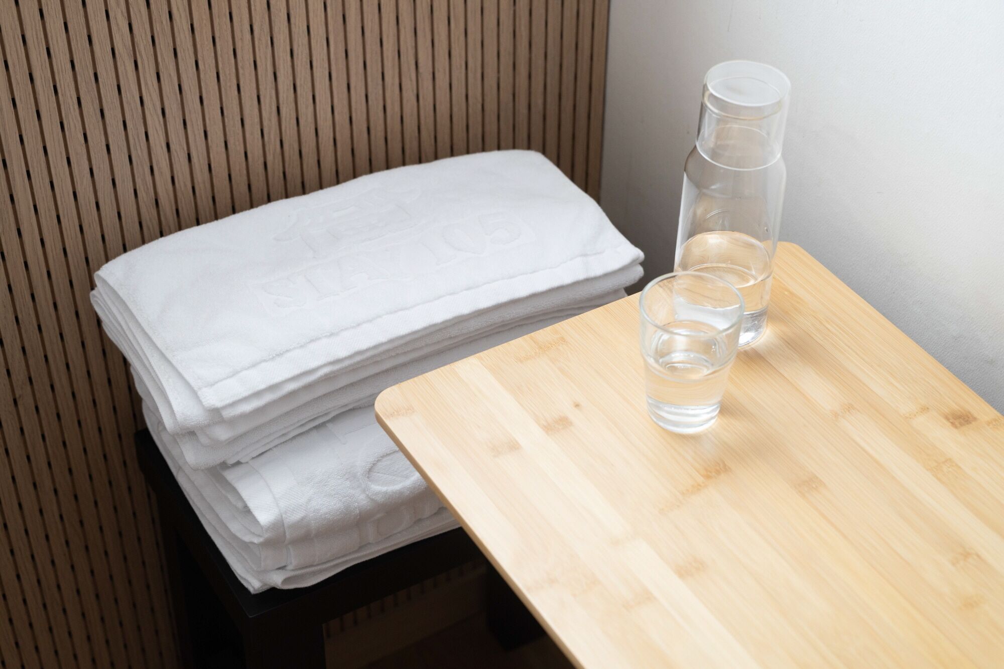 A neatly stacked set of white towels on a wooden chair, a clear glass bottle, and two glasses on a wooden table.