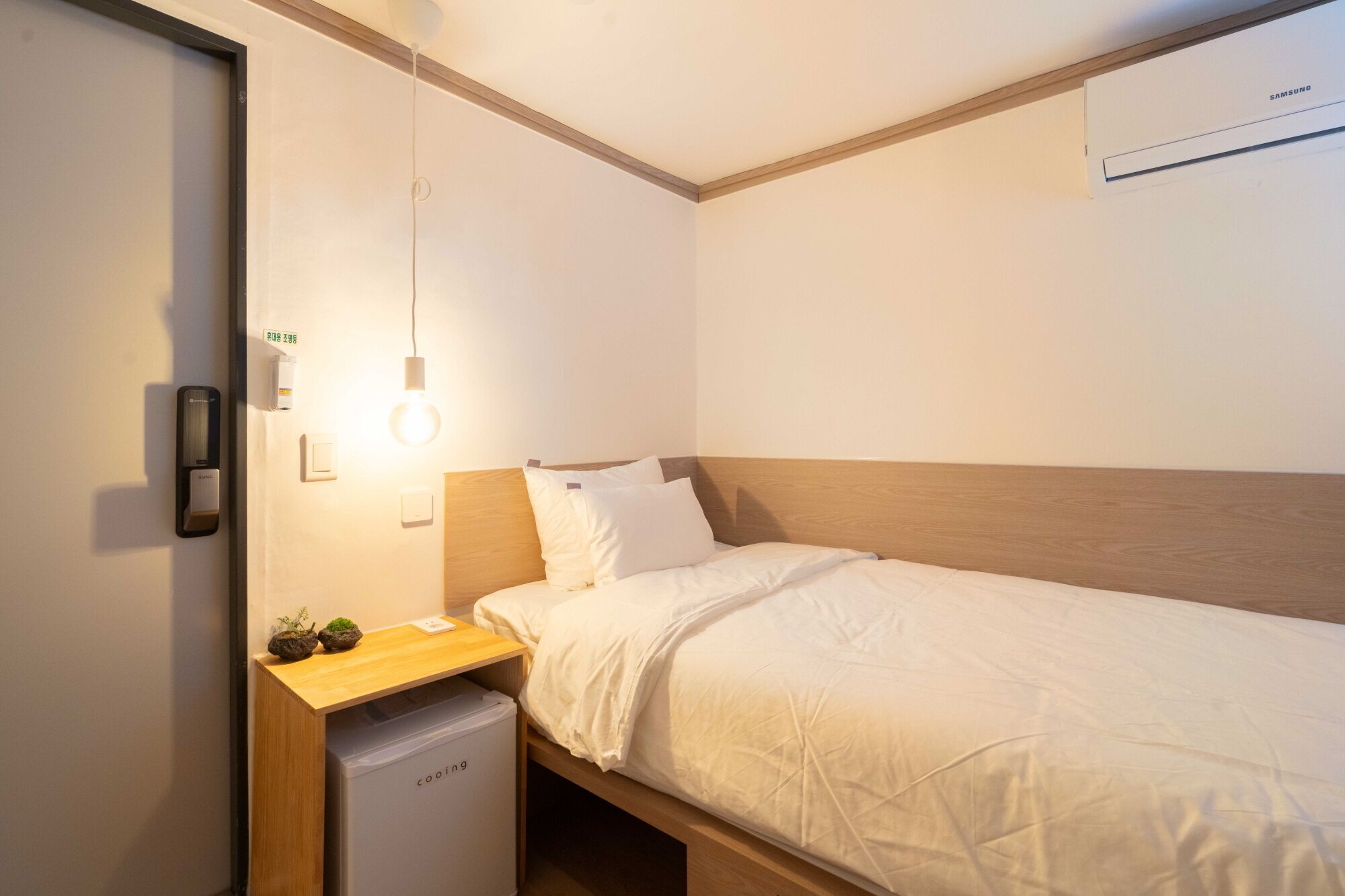 Une petite chambre à coucher, bien rangée, avec un lit simple, une table de chevet avec une lampe, un petit réfrigérateur et un climatiseur fixé au mur.