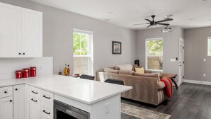 Una cucina moderna con mobili bianchi, un'isola centrale e forno integrato. C'è un divano beige con una coperta rossa e un ventilatore a soffitto.