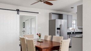 A dining area with a wooden table and six chairs, a ceiling fan, and a kitchen with grey cabinets and stainless steel appliances. - Austin Country Blues Home 3b/2b Close to Town, Zilker Park (Austin)