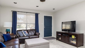 A living room with a brown leather sofa, a grey ottoman, a flat-screen TV on a wooden cabinet, and a blue door. - Austin Country Blues Home 3b/2b Close to Town, Zilker Park (Austin)