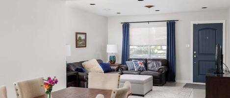 A modern living room with a dining area, a dark-colored sofa, a coffee table, and a large window with curtains.