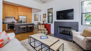 Eine moderne Küche mit Holzschränken, einem Edelstahlkühlschrank und einem eingebauten Backofen. Es gibt einen Essbereich mit einem Holztisch und Stühlen, sowie ein Wohnzimmer mit Kamin und einer Couch.