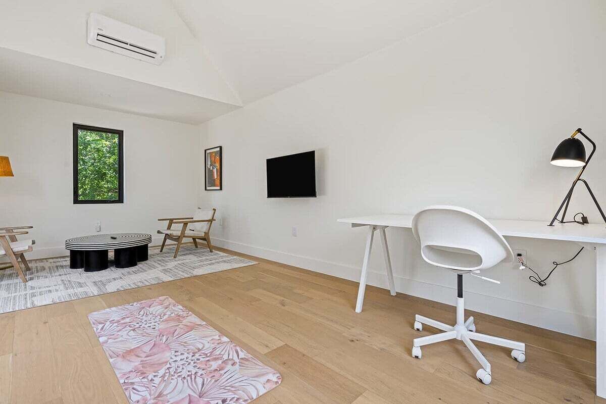 A modern, minimalistic room with a wooden floor, a white desk, a white office chair, a round table with chairs, a flat-screen TV mounted on the wall, and a window with a view of trees.