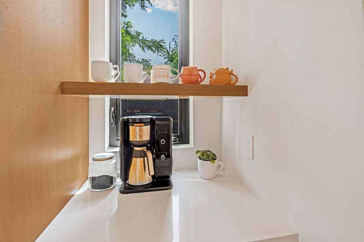 Ett köksutrymme med en kaffebryggare, en hylla med muggar och ett fönster med utsikt över grönområden.