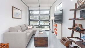 En moderne stue med en grå sofa, et træbord, et fjernsyn og et stort vindue med udsigt til en bil udenfor.