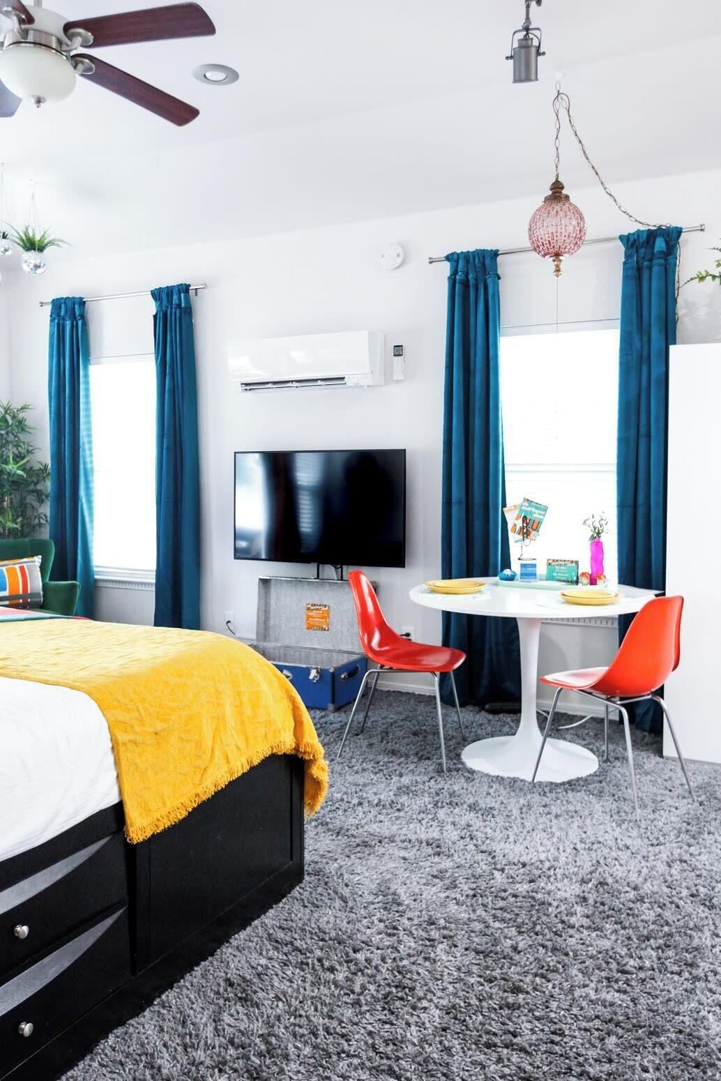 A modern hotel room with a bed, a TV, a dining table with chairs, and a ceiling fan.
