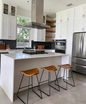 A modern kitchen with white cabinets, a central island with stools, stainless steel appliances, and a view of greenery outside. - 3br, Rooftop Deck, Walk In Shower - Nest (Austin)